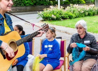 two primary aged school children and an older woman, seated, singing lead by a woman playing a guitar